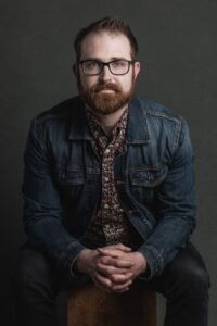 A person in a denim jacket and floral shirt sits on a wooden stool against a dark backdrop.