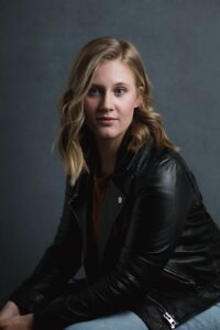 A woman in a black leather jacket sits against a dark backdrop.