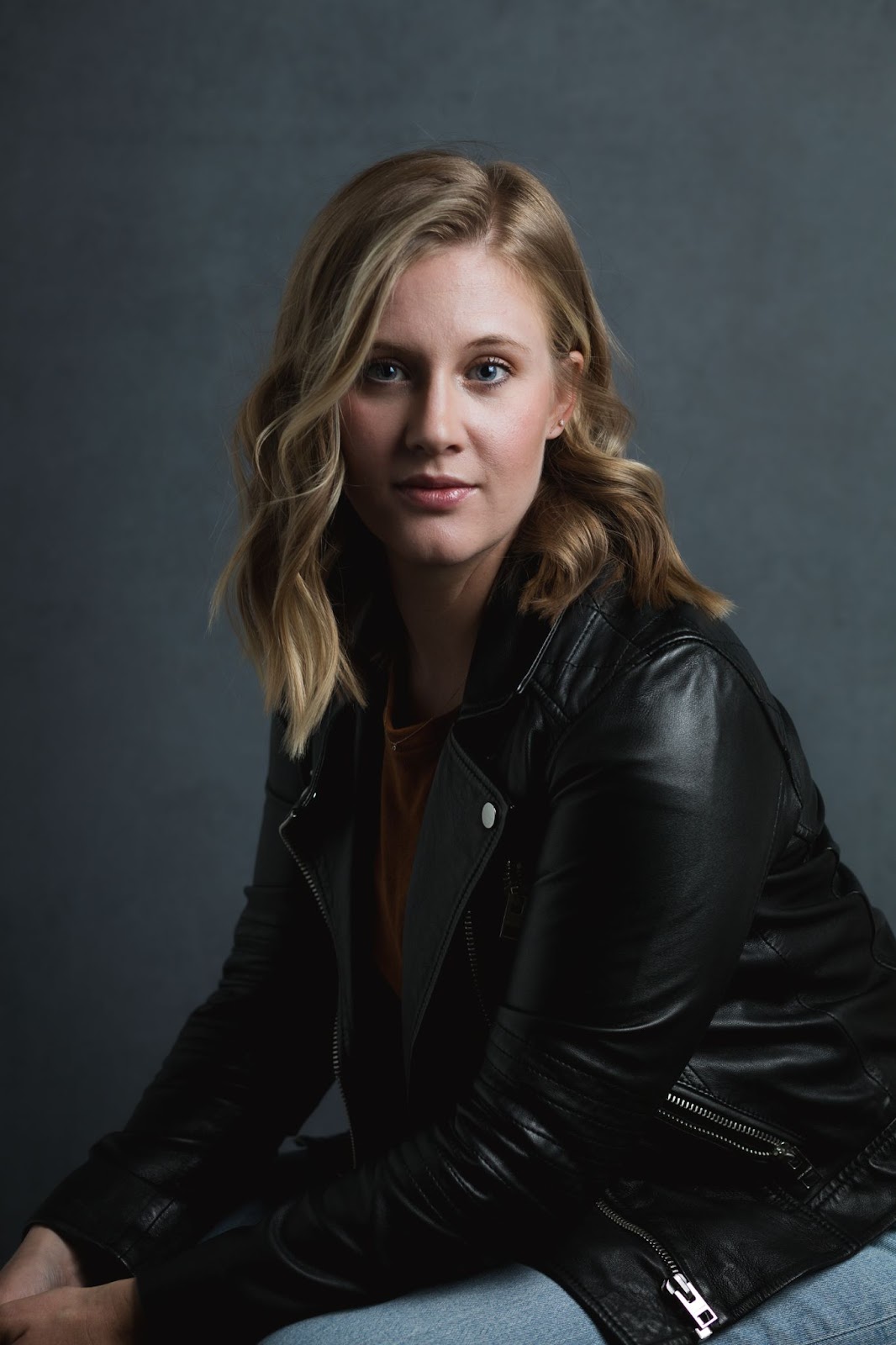 A woman in a black leather jacket sits against a dark backdrop.
