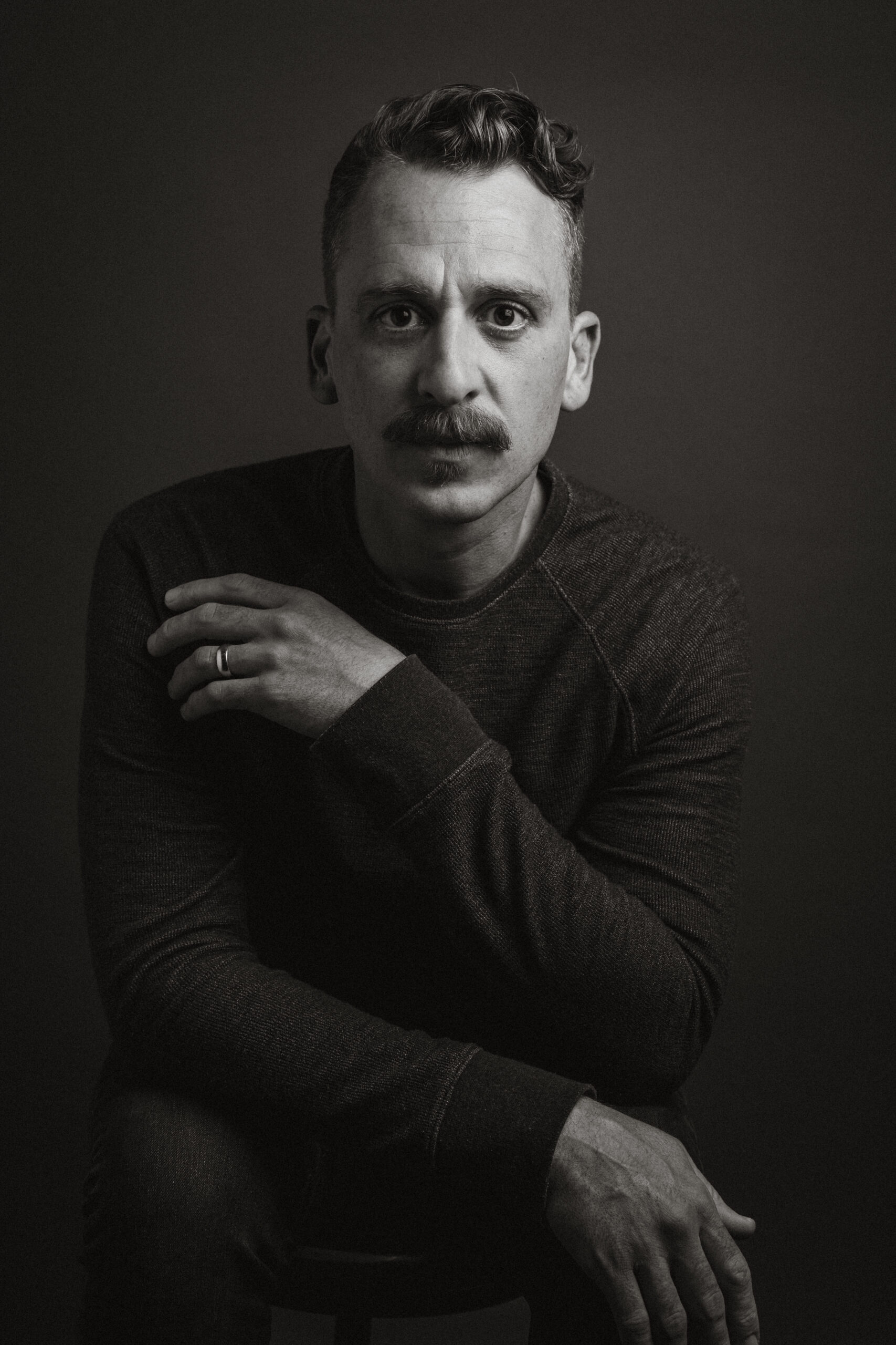 A man with a mustache is seated comfortably on a chair, exuding a relaxed demeanor.