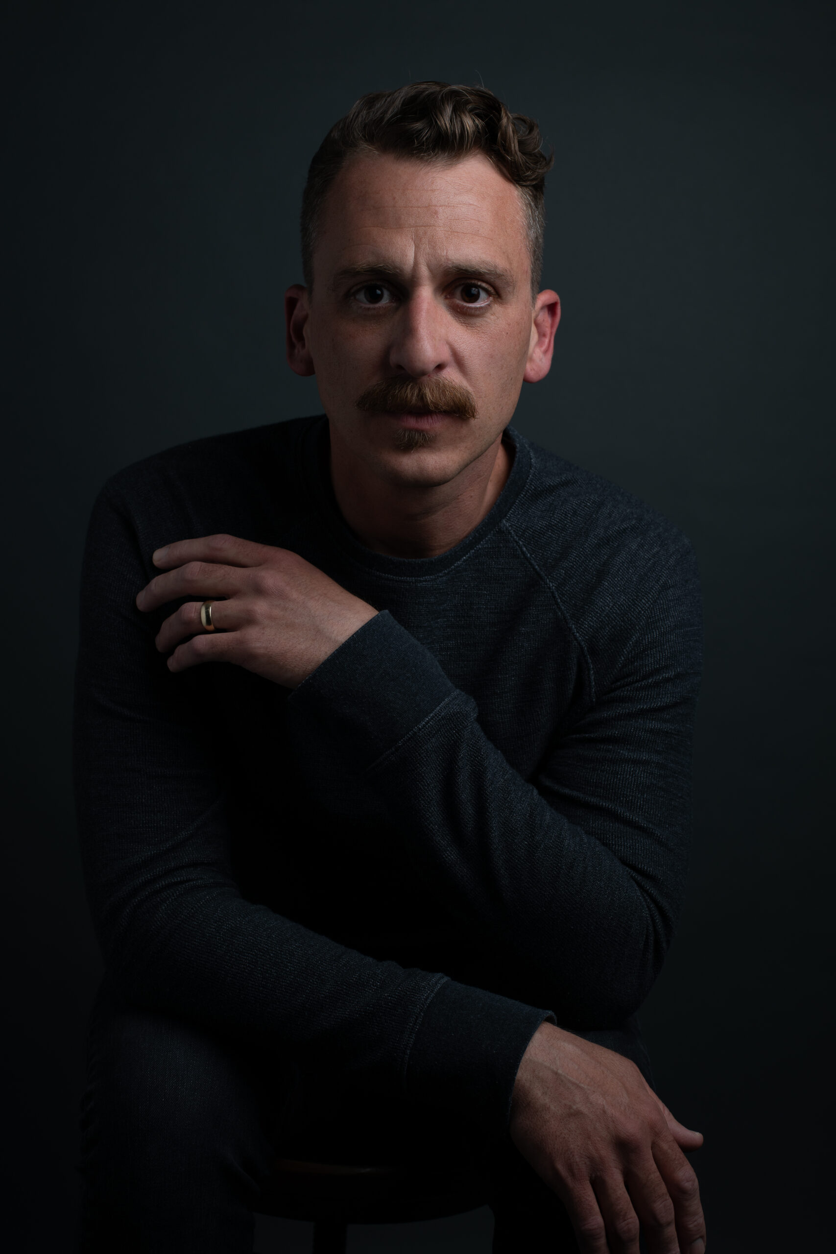 A man sporting a mustache sits in a chair.