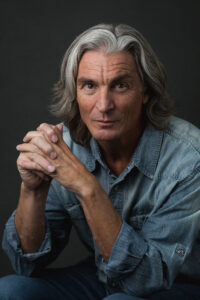 A man with long white hair wearing a blue shirt, standing confidently with a serene expression.