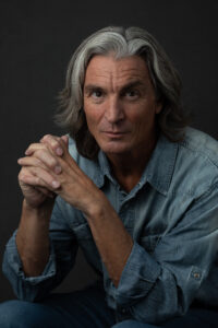 A man sporting long white hair and a blue shirt, presenting a tranquil and dignified appearance.