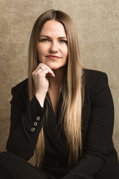 A woman in a black suit is seated on a chair, exuding professionalism and confidence in her posture.