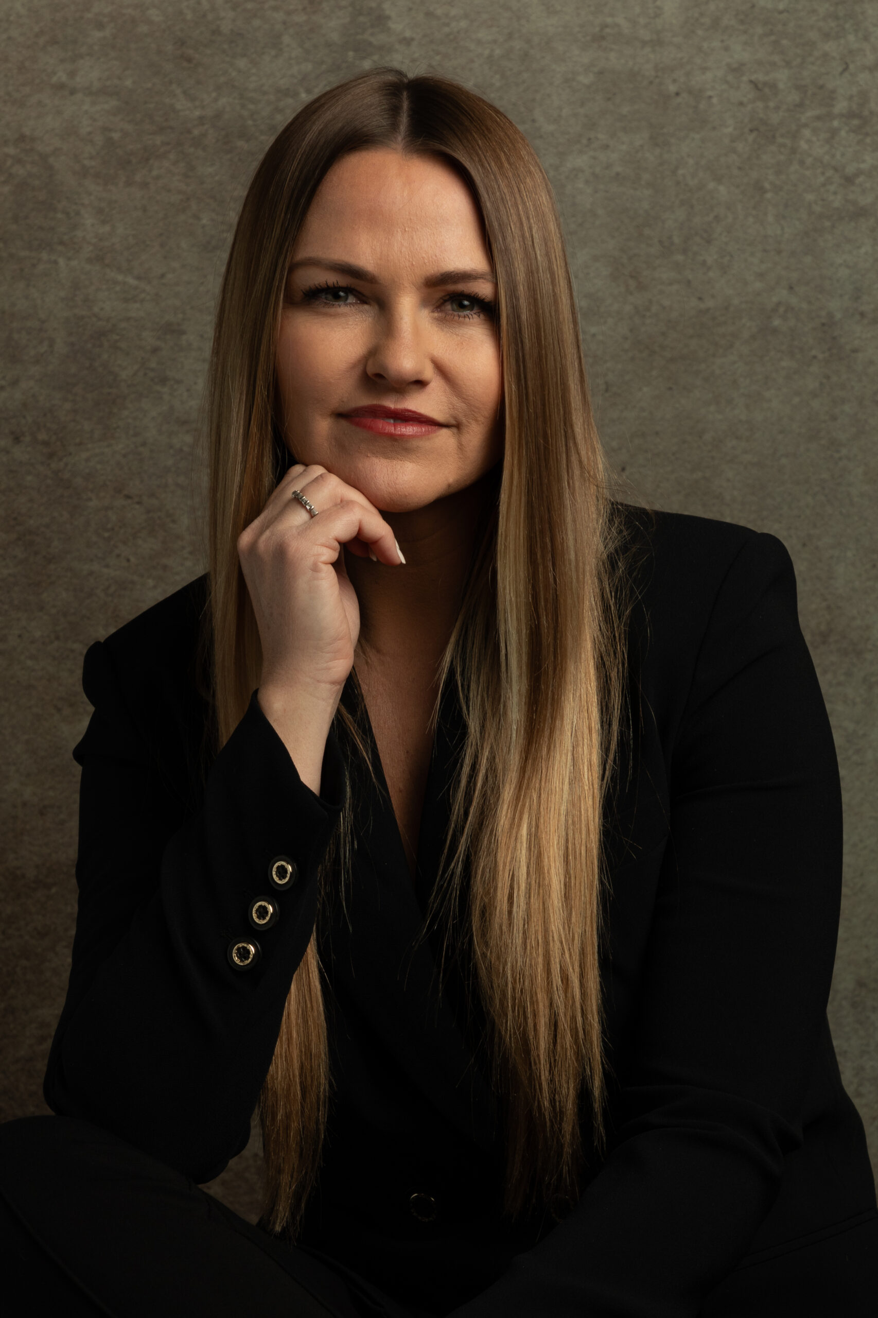 A woman dressed in a black suit sits elegantly on a chair, showcasing a poised and professional demeanor.