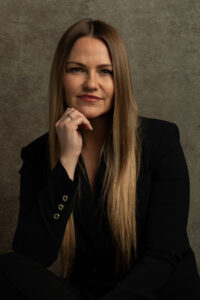 A woman dressed in a black suit sits elegantly on a chair, showcasing a poised and professional demeanor.