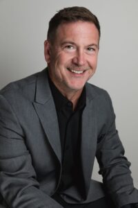 A smiling man in a suit and black shirt, captured in a professional corporate headshot setting.