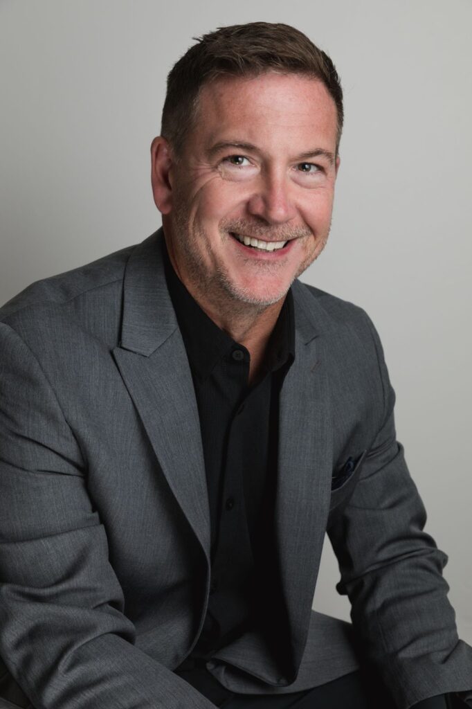 A smiling man in a suit and black shirt, captured in a professional corporate headshot setting.