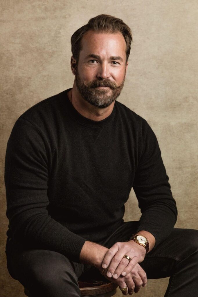 A bearded man in a black sweater is seated on a stool, exuding a relaxed and contemplative demeanor