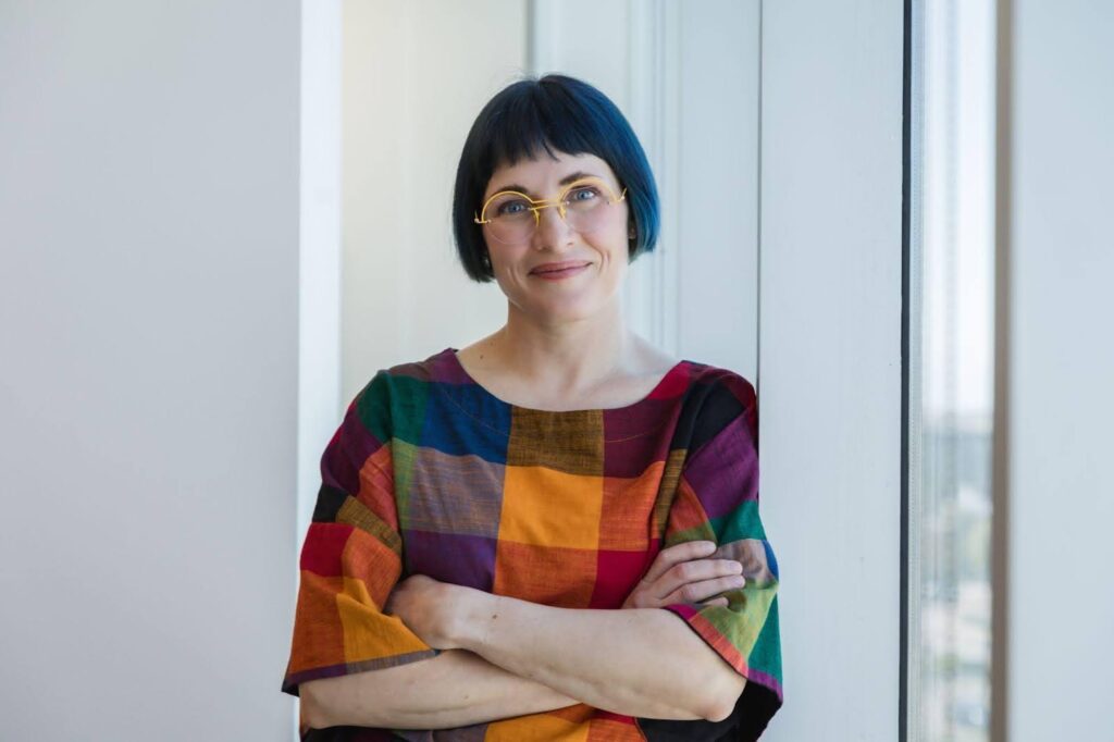 A woman with glasses and a colorful shirt poses by a window, radiating positivity