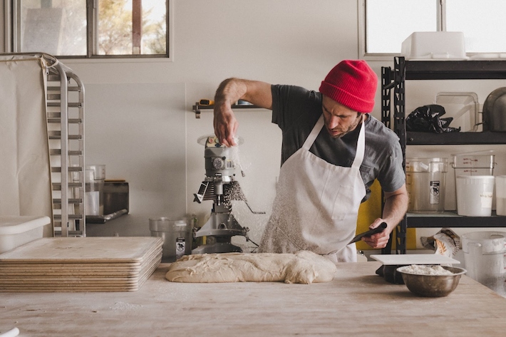 A baker in a red beanie and apron sprinkles flour over dough on a wooden countertop in a bright, organized bakery kitchen.