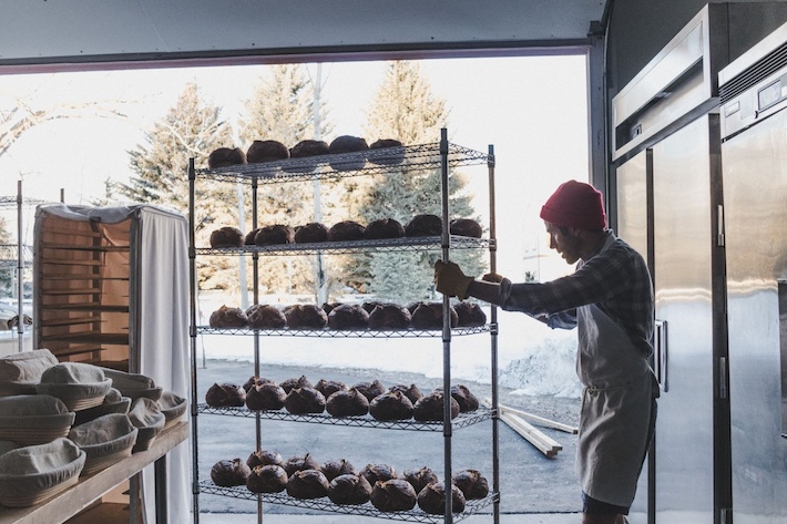 A baker wearing a red beanie pushes a metal rack filled with freshly baked loaves of bread in a sunlit bakery, with snowy trees visible outside