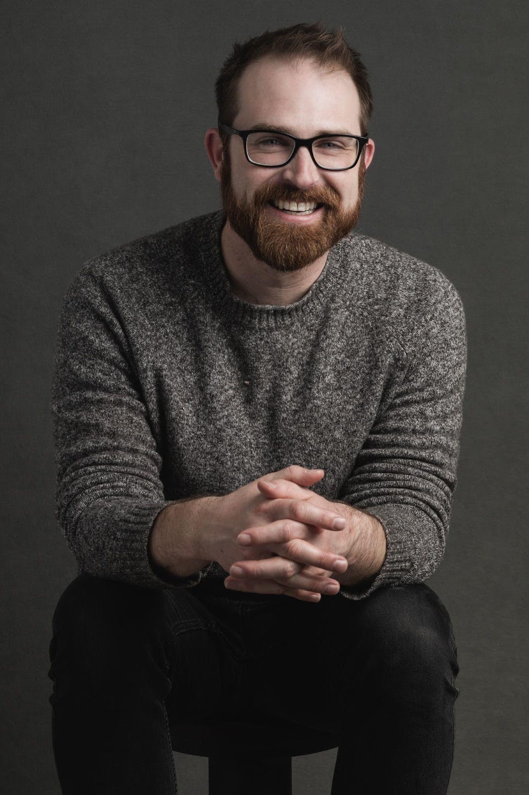 A bearded man wearing glasses sits on a stool, exuding a calm and contemplative demeanor