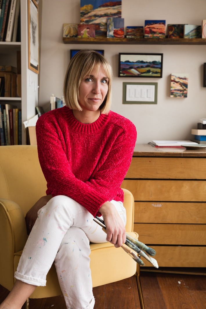 A woman in a red sweater sits in a yellow chair holding paintbrushes, surrounded by colorful artwork in a cozy studio.