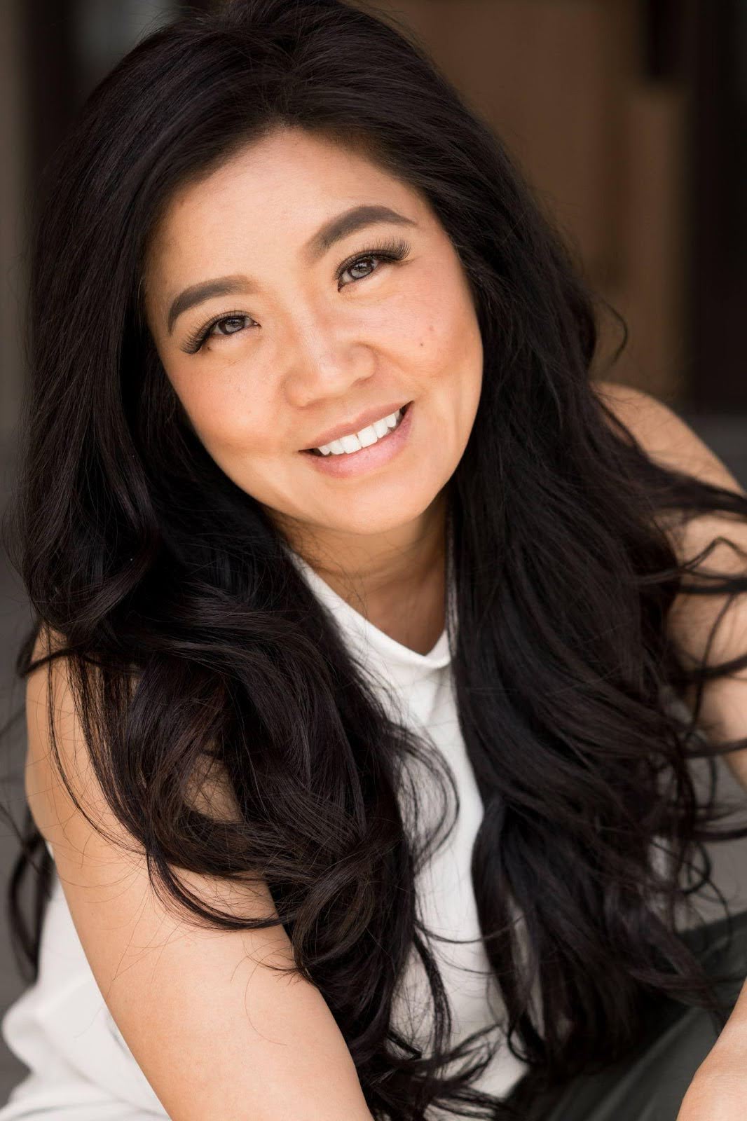 A woman with long black hair wearing a white shirt, standing confidently against a neutral background