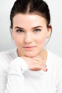 Close-up portrait of a woman with natural makeup, blue eyes, and gold jewelry, posing gracefully in a white top.