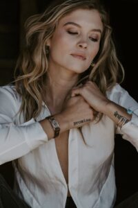 Woman with elegant makeup and meaningful wrist tattoos, posing thoughtfully in a white blouse.