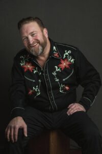 A bearded man in a cowboy shirt sits comfortably on a stool showcasing a casual and rustic style