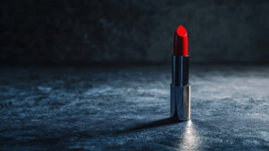 A vibrant red lipstick in a metallic tube stands upright on a textured dark surface. The soft lighting creates a dramatic, elegant atmosphere.