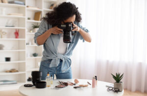 A woman in a denim shirt photographs makeup products on a white table. A lens, perfume, and a small plant are nearby. The room feels bright and creative.