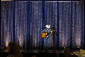 A musician in a cowboy hat plays guitar on a dimly lit stage with a sparkling curtain backdrop, creating a serene and intimate atmosphere.