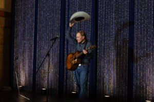 A man in a denim shirt and jeans stands on stage holding a guitar, waving a cowboy hat. Behind him, sparkling blue curtains add a festive tone.