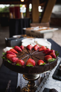 A decadent chocolate cake on a silver stand, topped with fresh strawberries and mint leaves. The elegant setting suggests a festive occasion.