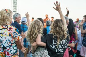 Back view of a crowd at an outdoor concert, featuring a woman with a child in her arms. The woman’s arm is raised, creating a lively, celebratory atmosphere.