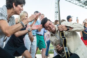 Guitarist energetically plays amid an enthusiastic crowd at an outdoor concert. People clap and cheer, creating a lively, joyful atmosphere.