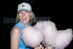 A smiling person in a white cap holds large pink cotton candy in both hands at night. The scene is joyful, with a sense of fun and excitement.