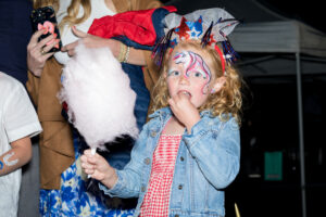 A young girl with face paint of a unicorn and festive headwear holds pink cotton candy. She wears a denim jacket and gingham dress, creating a playful, joyful scene.