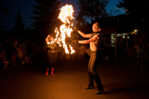 Two performers engage in a nighttime fire show. One holds a large, fiery prop, while the other gestures dramatically. A captivated crowd watches.