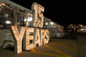 Illuminated "15 Years" sign with glowing bulbs stands outside a lit tent at night, conveying a celebratory and festive atmosphere.