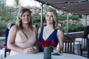 Two women in evening dresses pose at an outdoor event under string lights. They stand by a table with red roses, smiling and holding drinks.