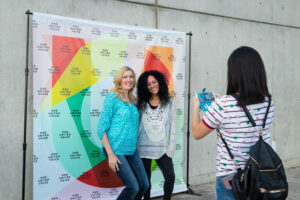 Two women pose happily for a photo in front of a colorful "One United Voice" backdrop. A third person takes their picture on a phone, conveying a joyful and lively atmosphere.