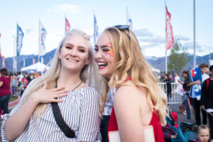 Two women with face paint smile at an outdoor event, surrounded by flags and a crowd against a mountain backdrop. The scene is joyful and lively.