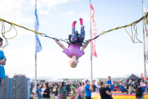 A child in a purple shirt and black pants joyfully bounces upside down on a bungee trampoline at a lively outdoor event, with crowds and flags in the background.
