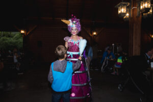 A child in a blue hoodie interacts with a woman in a colorful, candy-themed costume sporting a pink dress and elaborate headpiece inside a dimly lit hall.