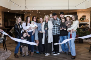 A group of people, including adults and children, happily gather inside a store for a ribbon-cutting ceremony. A woman in the center uses large scissors to cut the ribbon, while others smile and celebrate the moment. The space has a comfortable, inviting ambiance.