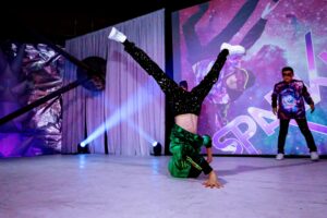 A breakdancer in a green outfit performs an inverted pose on the floor, legs in a V shape. Spectator in galaxy print outfit watches. Dynamic stage lighting.