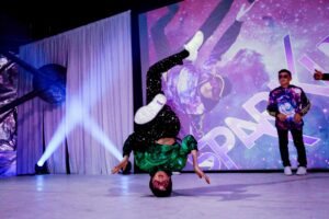 Breakdancer performing a headstand, wearing a vibrant outfit, in front of a cosmic-themed backdrop with the word "SPARKLE." Stage lights create a dynamic atmosphere.