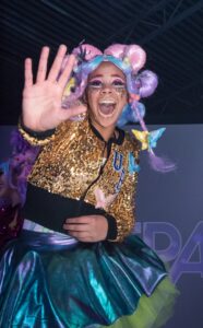 Person with colorful, whimsical makeup and attire looks joyous, wearing a golden sequin jacket and metallic skirt, waving energetically at the camera.
