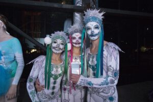 Three people in elaborate costumes with colorful skeleton face paint, adorned with sparkling gems, convey a festive and theatrical mood.