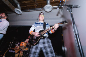 Man playing electric guitar on stage, wearing striped shirt, plaid pants, and headband. Energetic performance with drummer and microphone nearby. Industrial ceiling.