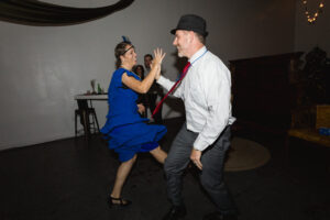 A woman in a vibrant blue dress and a man in a white shirt and hat are joyfully swing dancing. The setting appears festive and lively.